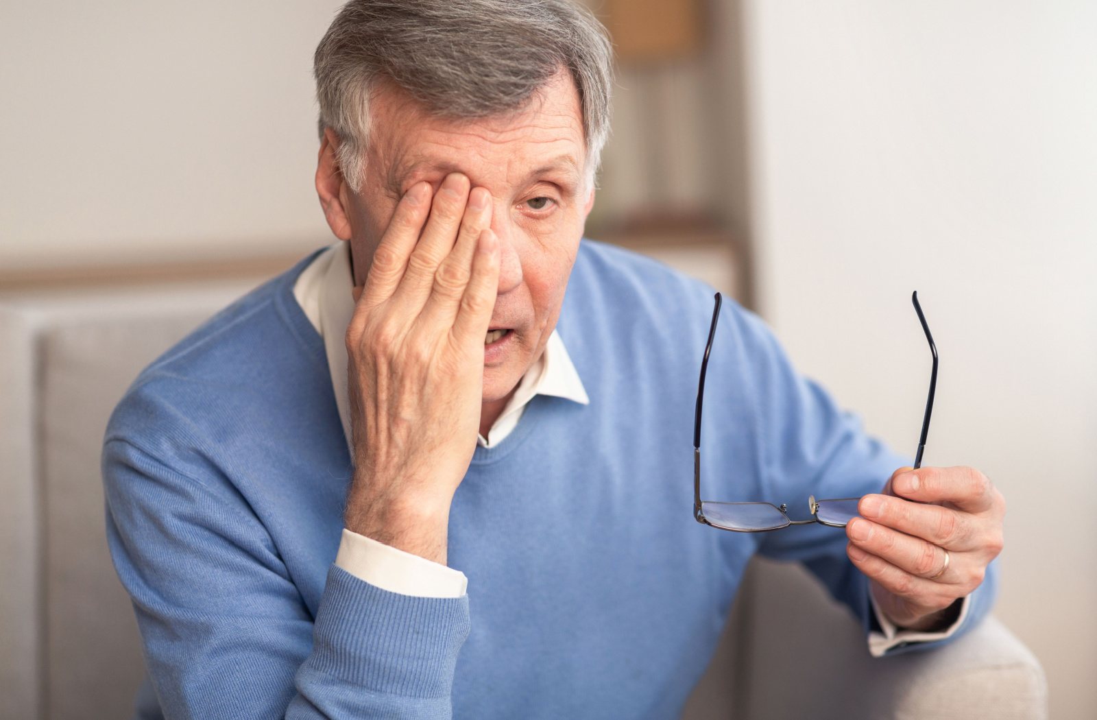 An older adult man sitting on a couch and holding his glasses in his left hand as he rubs his right eye with his other hand.