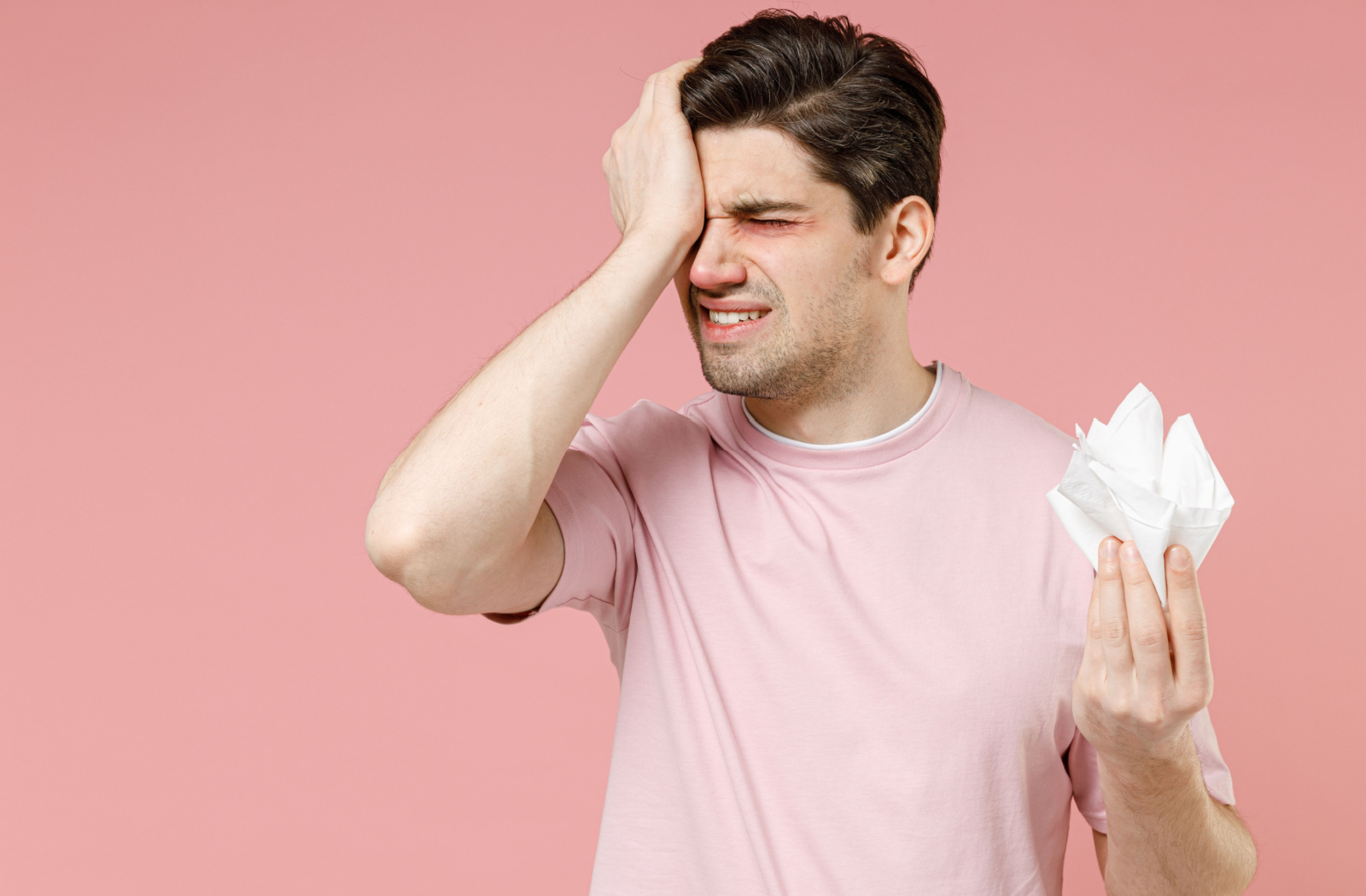 A young man holding a tissue in his left hand and rubbing his right eye in irritation with his right hand