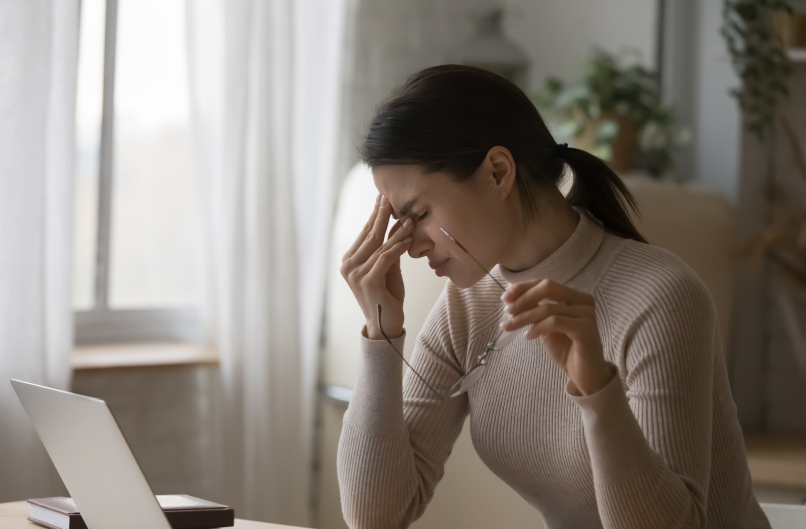 A woman sitting at her computer holding her glasses and rubbing hey eyes and forehead.
